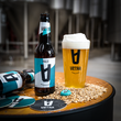 Load image into Gallery viewer, Vetra Pale Gluten Free West Coast IPA. Sul un tavolo di legno rotondo sono disposti: una bottiglia di birra Vetra Pale, un bicchiere pieno di birra chiara con schiuma abbondante e il logo Vetra, dei sottobicchieri con il logo Vetra, dei chicchi di malto sparsi sulla superficie. La birra nel bicchiere ha un colore dorato brillante con una generosa corona di schiuma bianca.