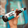 Load image into Gallery viewer, Mano che tiene una bottiglia di Vetra Pale, birra artigianale senza glutine West Coast IPA. Lo sfondo presenta toni sfocati di blu e verde che creano un contrasto vivace con il design della bottiglia. La scena cattura l'essenza urbana e fresca della birra #birradistrada, ideale per chi cerca una IPA gluten free con personalità.